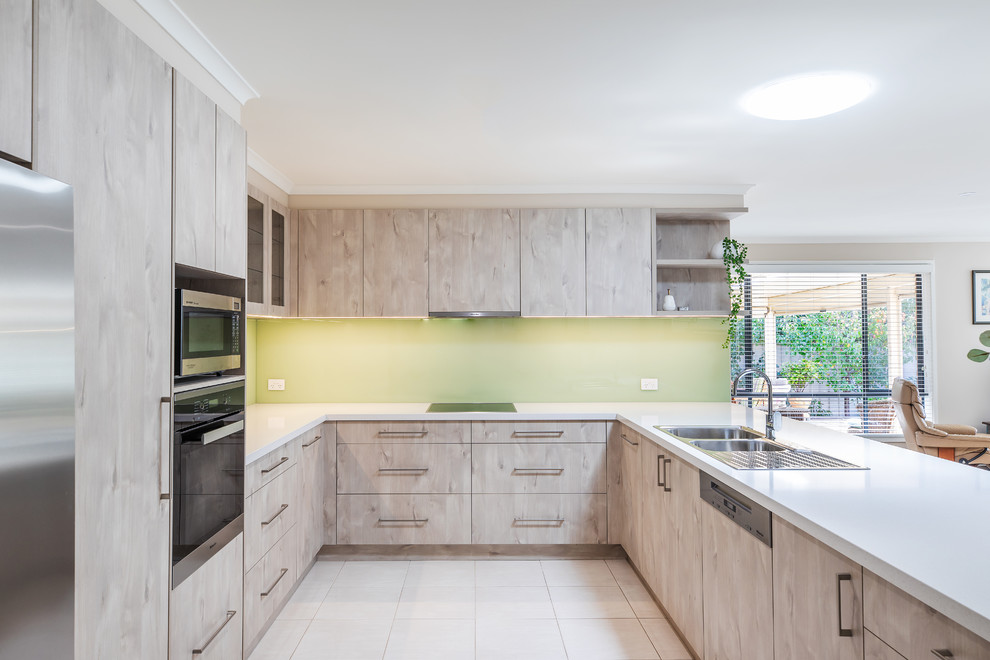 Inspiration for a mid-sized modern u-shaped kitchen pantry in Adelaide with a double-bowl sink, flat-panel cabinets, light wood cabinets, quartz benchtops, green splashback, glass sheet splashback, stainless steel appliances, porcelain floors, a peninsula, white floor and white benchtop.