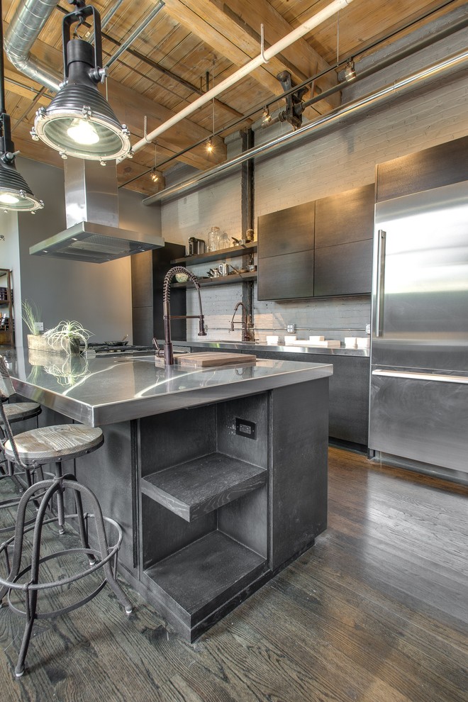 This is an example of a mid-sized industrial galley open plan kitchen in Chicago with a double-bowl sink, flat-panel cabinets, grey cabinets, stainless steel benchtops, grey splashback, stainless steel appliances, light hardwood floors and with island.