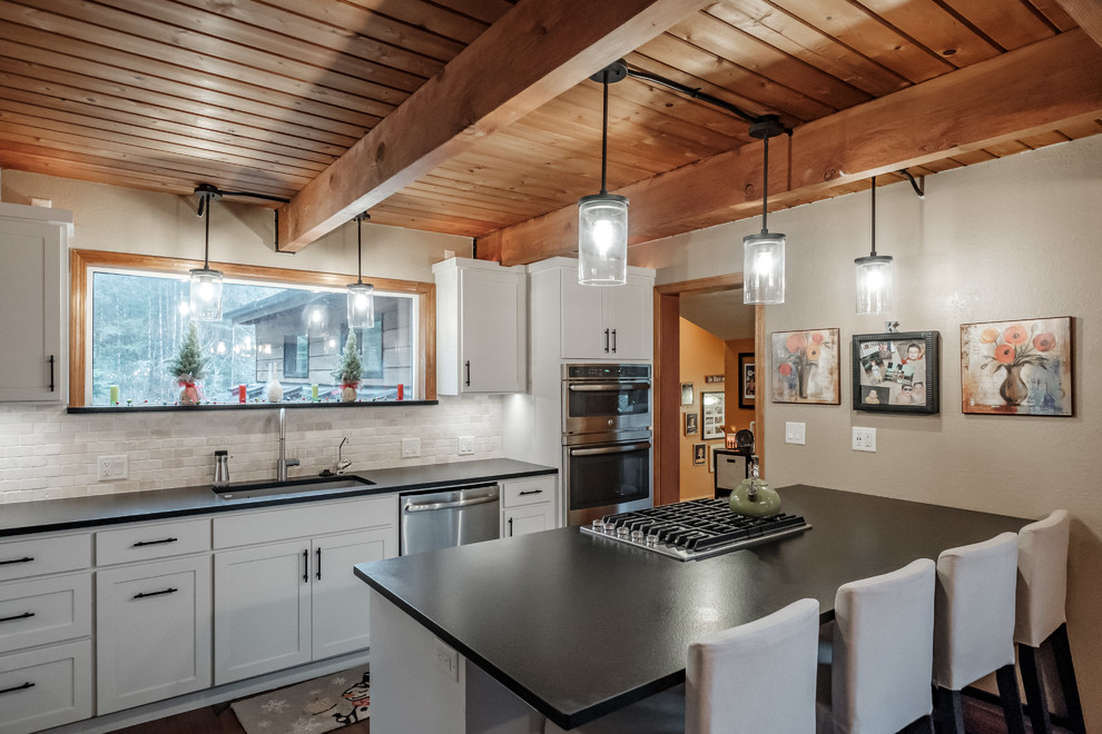 This is an example of a large country l-shaped eat-in kitchen in Portland with a single-bowl sink, shaker cabinets, white cabinets, granite benchtops, white splashback, stone tile splashback, stainless steel appliances, dark hardwood floors, with island, brown floor and black benchtop.