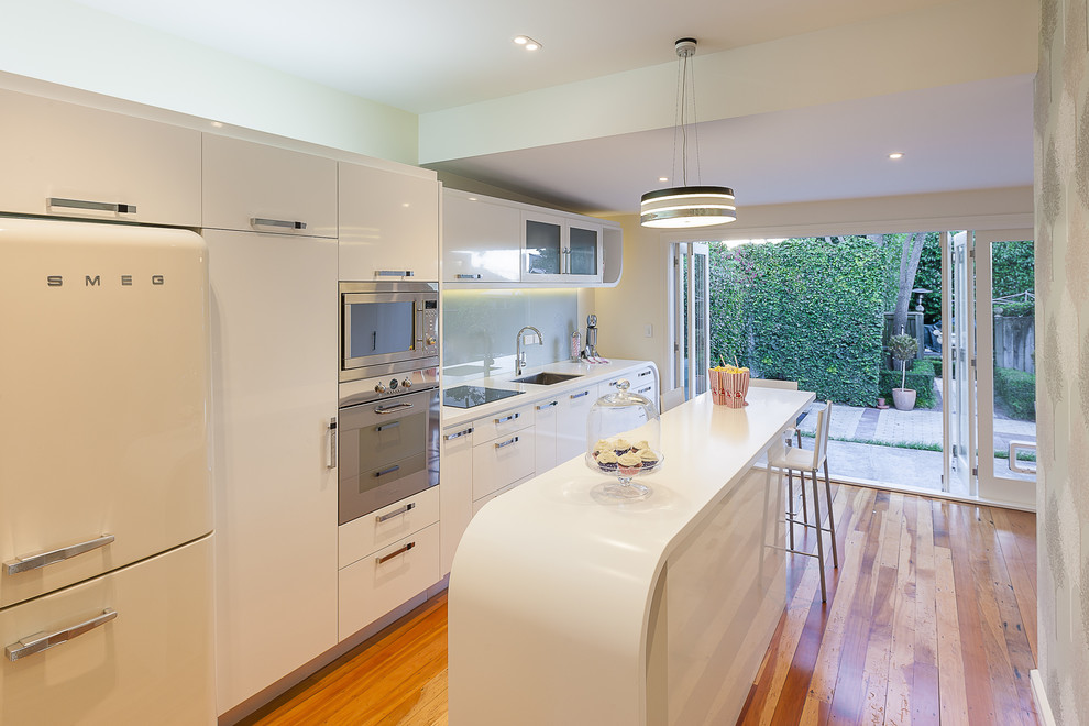Modern single-wall kitchen in Auckland with an undermount sink, flat-panel cabinets, white cabinets, glass sheet splashback and white appliances.