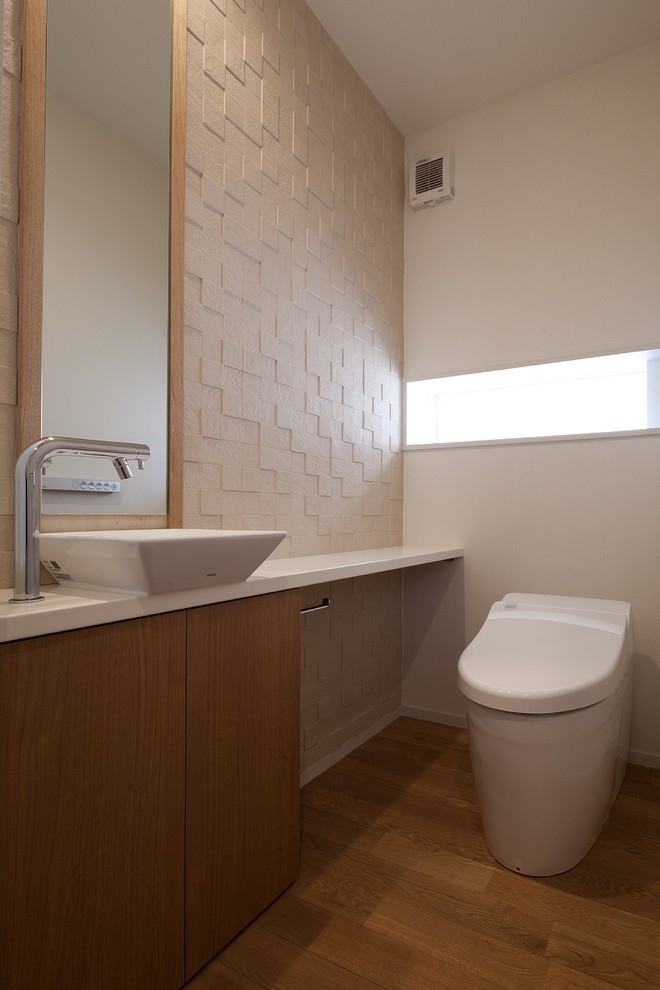 Cette image montre un WC et toilettes nordique avec des portes de placard beiges, un mur beige, un sol en bois brun, un plan de toilette en surface solide, un sol marron et un plan de toilette blanc.
