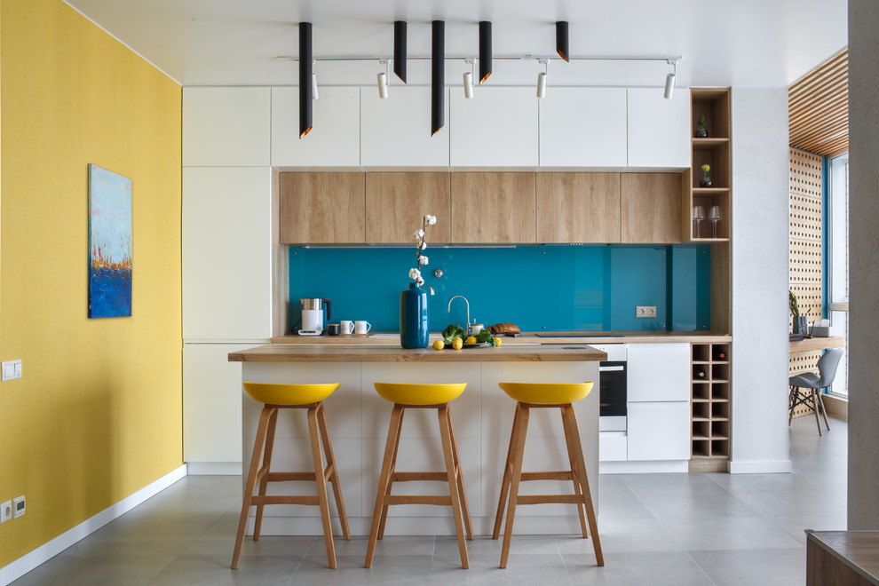 Contemporary single-wall kitchen in Other with a drop-in sink, flat-panel cabinets, white cabinets, wood benchtops, blue splashback, glass sheet splashback, stainless steel appliances, with island, grey floor and brown benchtop.