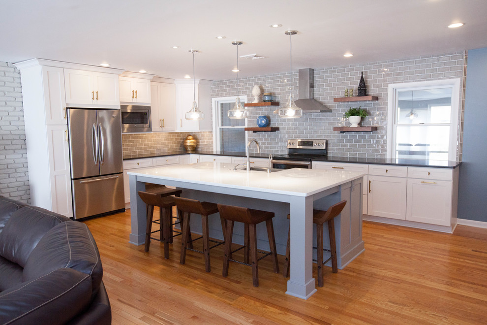 Large contemporary l-shaped open plan kitchen in Philadelphia with a farmhouse sink, shaker cabinets, white cabinets, quartzite benchtops, grey splashback, subway tile splashback, stainless steel appliances, medium hardwood floors and with island.