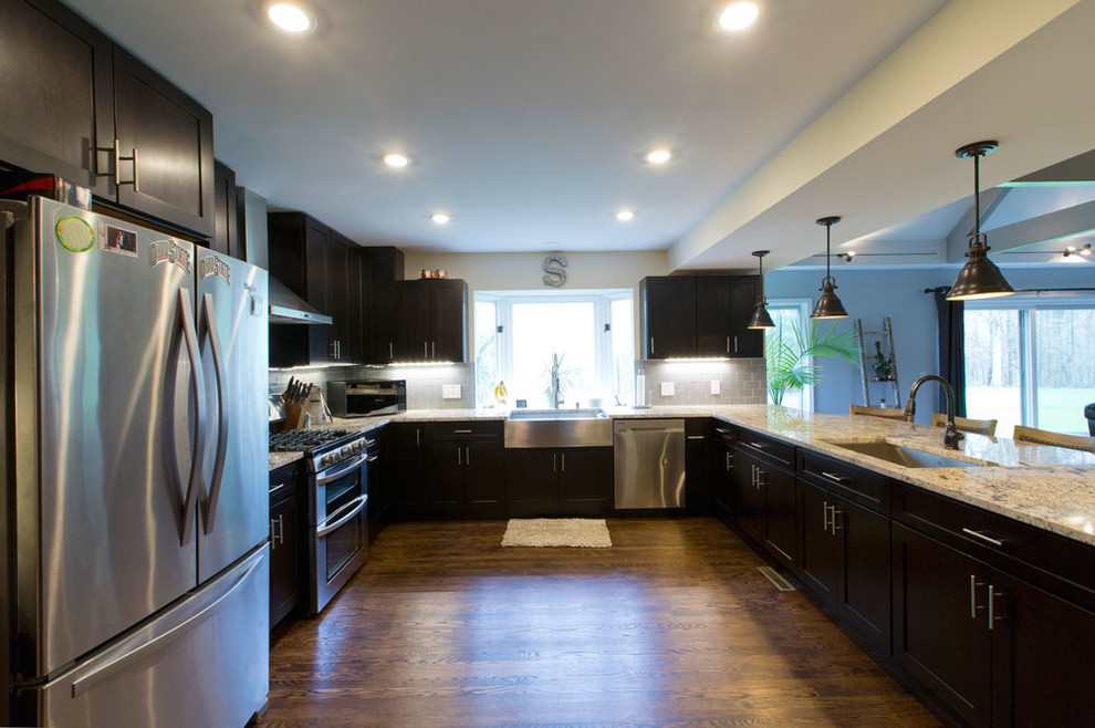 Large transitional u-shaped light wood floor and brown floor enclosed kitchen photo in Cleveland with a farmhouse sink, shaker cabinets, dark wood cabinets, granite countertops, gray backsplash, glass tile backsplash, stainless steel appliances, a peninsula and beige countertops