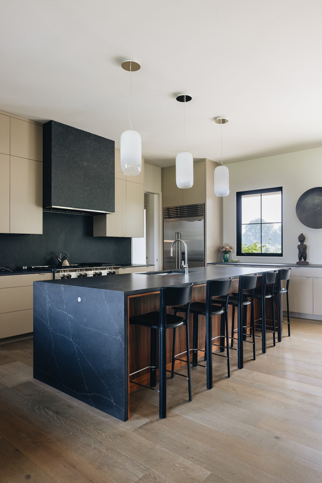 Design ideas for a large modern u-shaped open plan kitchen in Grand Rapids with a submerged sink, flat-panel cabinets, beige cabinets, marble worktops, black splashback, marble splashback, stainless steel appliances, light hardwood flooring, an island, brown floors and black worktops.