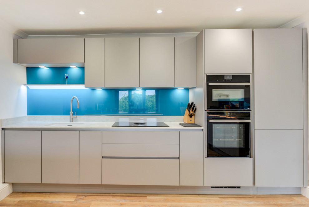 Immagine di una cucina moderna di medie dimensioni con lavello sottopiano, ante lisce, ante grigie, top in quarzite, paraspruzzi blu, paraspruzzi con lastra di vetro, elettrodomestici da incasso, pavimento in vinile, pavimento marrone e top bianco