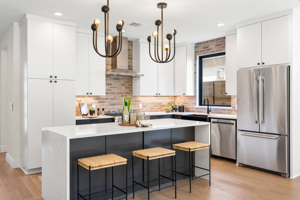 Photo of a traditional l-shaped open plan kitchen in Austin with a belfast sink, flat-panel cabinets, white cabinets, brick splashback, stainless steel appliances, medium hardwood flooring, an island and white worktops.