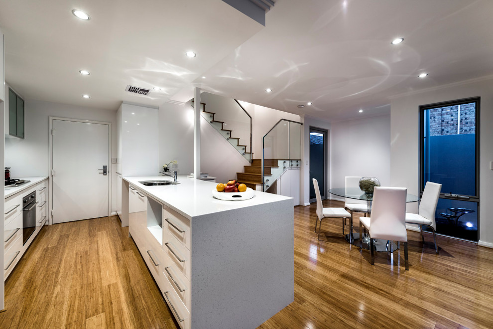 Photo of a contemporary eat-in kitchen in Perth with a double-bowl sink, white cabinets, quartz benchtops, metallic splashback, glass sheet splashback, bamboo floors, with island, brown floor and white benchtop.