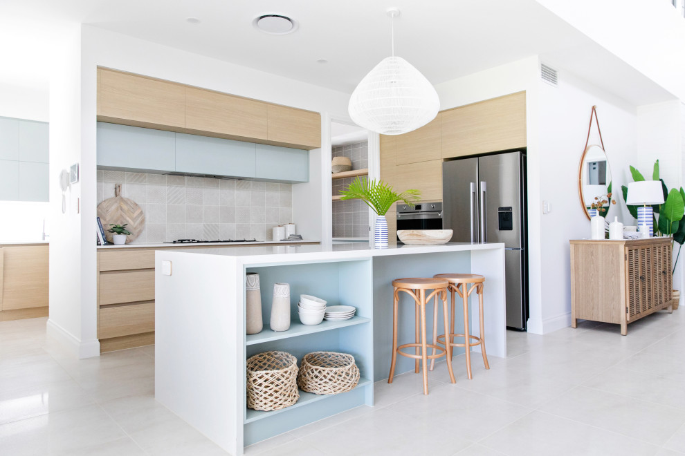 Inspiration for a mid-sized beach style galley open plan kitchen in Gold Coast - Tweed with an undermount sink, flat-panel cabinets, light wood cabinets, quartz benchtops, beige splashback, ceramic splashback, stainless steel appliances, ceramic floors, with island, white floor and white benchtop.