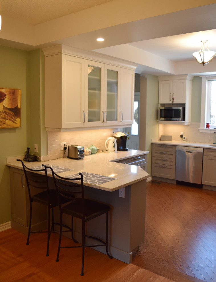 Example of a mid-sized transitional l-shaped medium tone wood floor eat-in kitchen design in Toronto with an undermount sink, shaker cabinets, white cabinets, quartz countertops, white backsplash, subway tile backsplash, stainless steel appliances and a peninsula