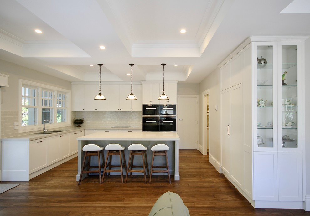 Design ideas for a large transitional l-shaped eat-in kitchen in Sydney with a drop-in sink, shaker cabinets, white cabinets, quartz benchtops, white splashback, porcelain splashback, black appliances, medium hardwood floors, with island and brown floor.