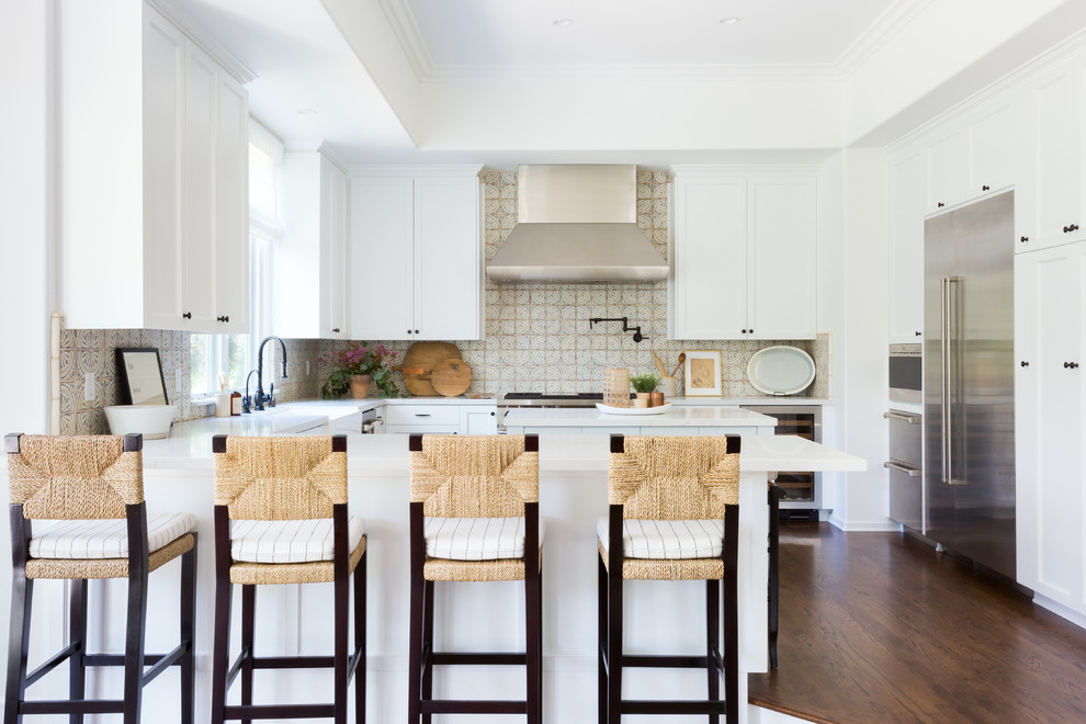 This is an example of a beach style u-shaped kitchen in Los Angeles with a farmhouse sink, shaker cabinets, white cabinets, multi-coloured splashback, stainless steel appliances, dark hardwood floors, a peninsula, brown floor and white benchtop.