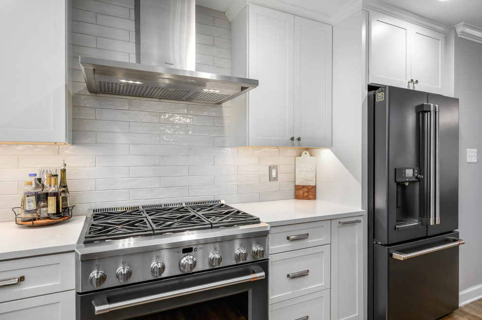 Photo of a classic l-shaped kitchen in DC Metro with a belfast sink, shaker cabinets, white cabinets, engineered stone countertops, white splashback, ceramic splashback, medium hardwood flooring, an island, brown floors and white worktops.