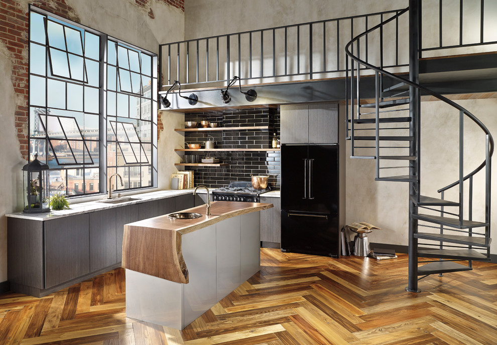 Design ideas for a mid-sized industrial l-shaped open plan kitchen in San Francisco with an undermount sink, flat-panel cabinets, grey cabinets, wood benchtops, black splashback, subway tile splashback, black appliances, medium hardwood floors and with island.
