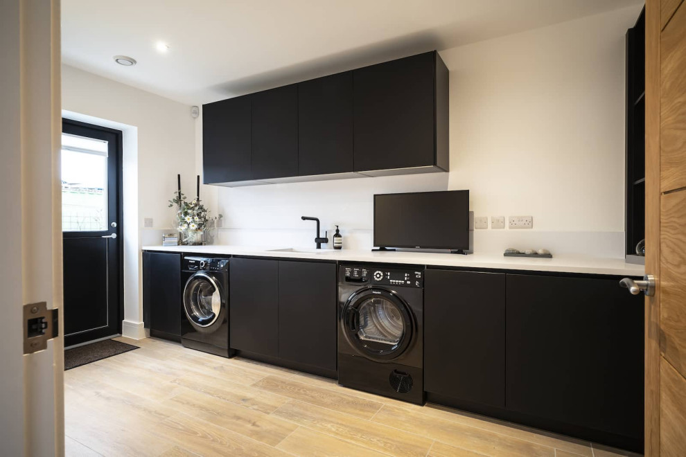 Photo of a large scandinavian l-shaped laundry room in Oxfordshire with flat-panel cabinets, black cabinets, quartzite benchtops, white splashback, light hardwood floors, brown floor and yellow benchtop.