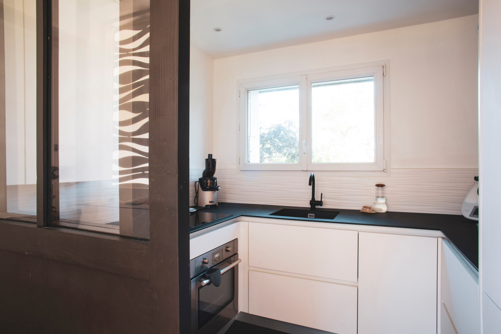 This is an example of a mid-sized beach style u-shaped separate kitchen in Other with an undermount sink, flat-panel cabinets, white cabinets, solid surface benchtops, white splashback, ceramic splashback, stainless steel appliances, terra-cotta floors, black floor and black benchtop.