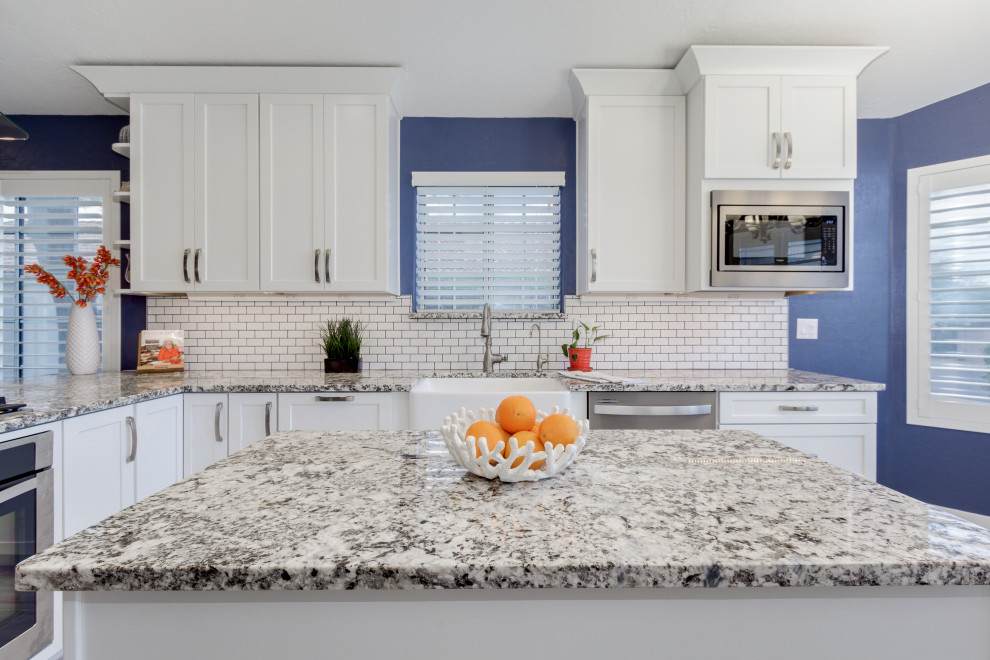 Example of a beach style porcelain tile and gray floor kitchen design in Other with a farmhouse sink, shaker cabinets, white cabinets, granite countertops, white backsplash, subway tile backsplash, stainless steel appliances, an island and multicolored countertops