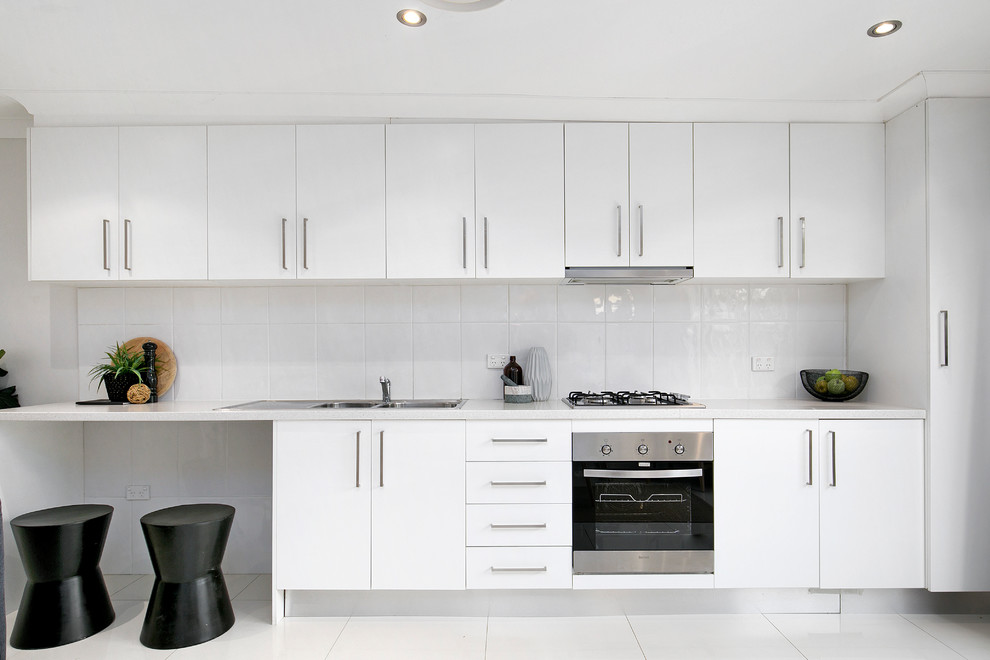 Photo of a contemporary single-wall kitchen in Sydney with a double-bowl sink, flat-panel cabinets, white cabinets, quartz benchtops, white splashback, ceramic splashback, stainless steel appliances, ceramic floors, no island, white floor and white benchtop.