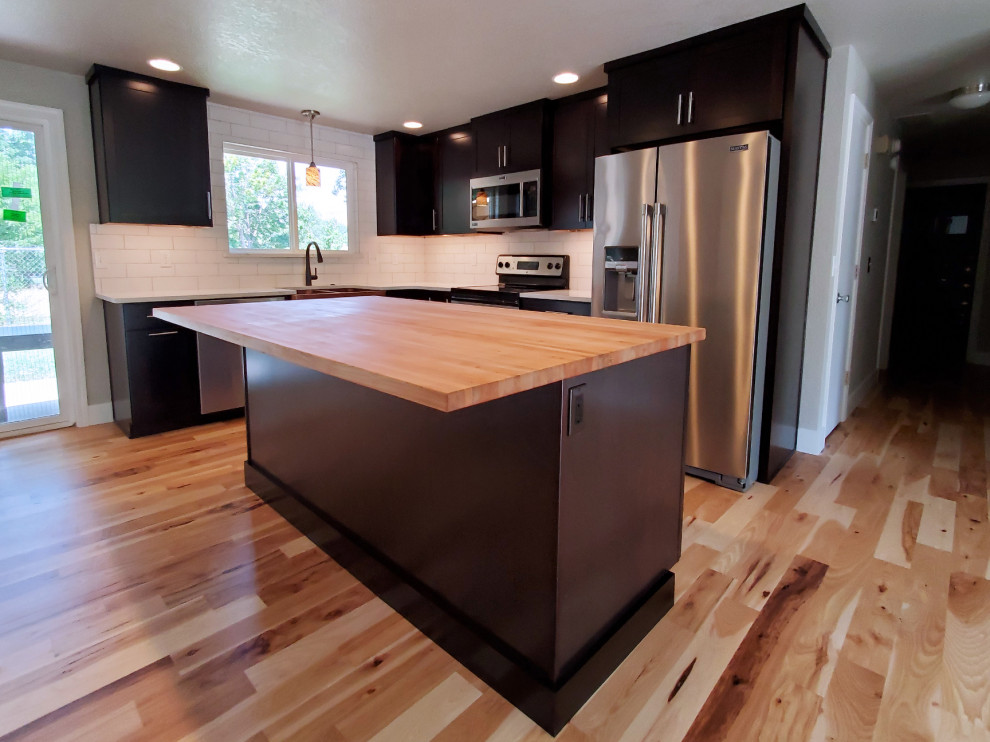 Inspiration for a mid-sized transitional l-shaped medium tone wood floor eat-in kitchen remodel in Boise with a farmhouse sink, shaker cabinets, black cabinets, quartz countertops, white backsplash, porcelain backsplash, stainless steel appliances, an island and white countertops