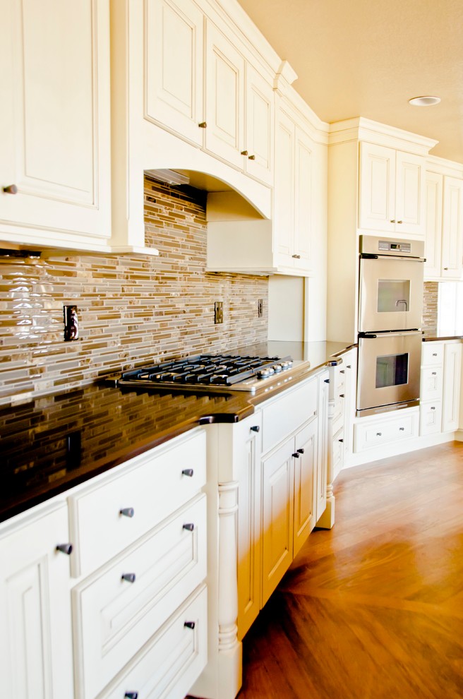 Large transitional l-shaped medium tone wood floor kitchen photo in Denver with white cabinets, granite countertops, black backsplash, glass tile backsplash, stainless steel appliances, an island, an undermount sink, raised-panel cabinets and white countertops