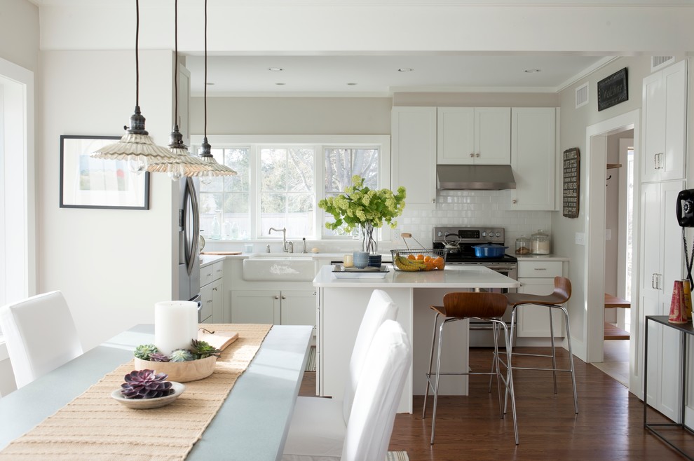 Photo of a mid-sized beach style l-shaped eat-in kitchen in New York with a farmhouse sink, shaker cabinets, white cabinets, white splashback, ceramic splashback, with island, stainless steel appliances, solid surface benchtops, dark hardwood floors and brown floor.