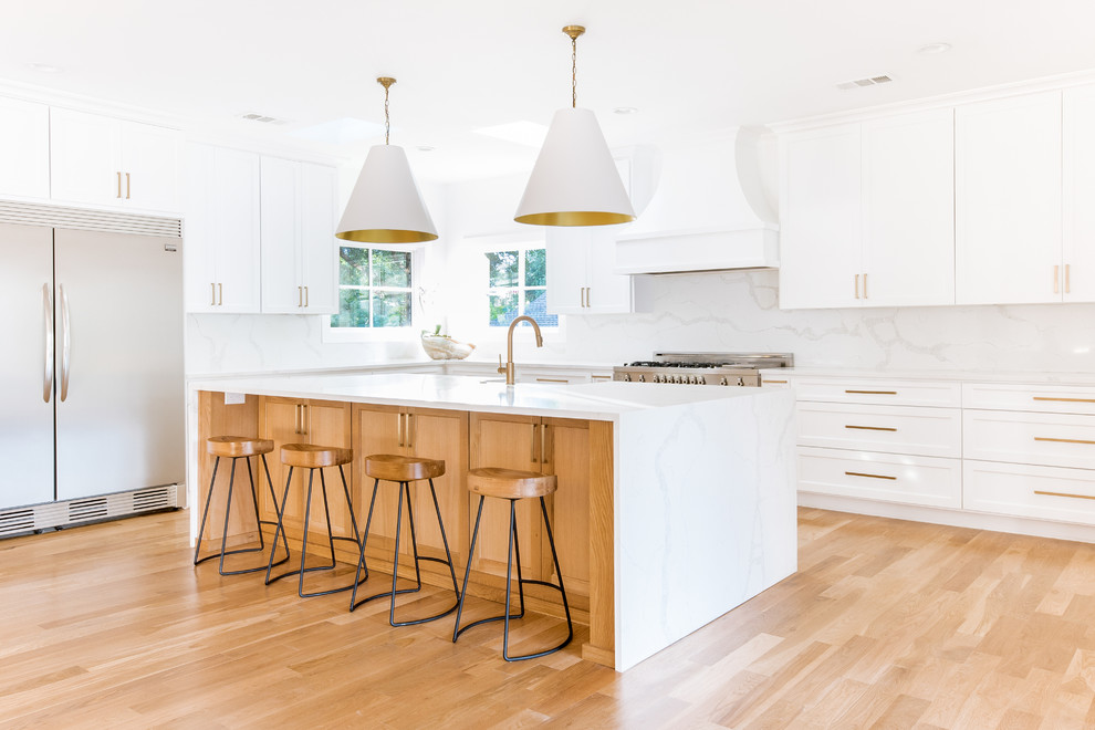 This is an example of a large transitional eat-in kitchen in Dallas with a farmhouse sink, shaker cabinets, white cabinets, quartz benchtops, white splashback, stone slab splashback, stainless steel appliances, light hardwood floors, with island and white benchtop.