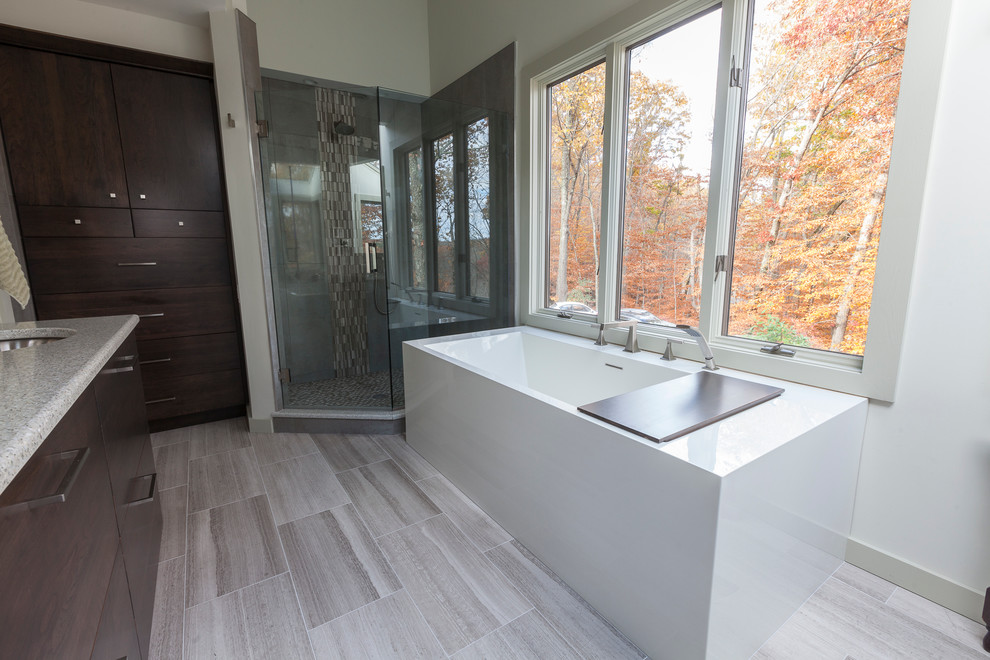 Inspiration for a large modern master bathroom in New York with flat-panel cabinets, brown cabinets, a freestanding tub, a corner shower, a two-piece toilet, gray tile, porcelain tile, grey walls, travertine floors, an undermount sink, recycled glass benchtops, grey floor and a hinged shower door.