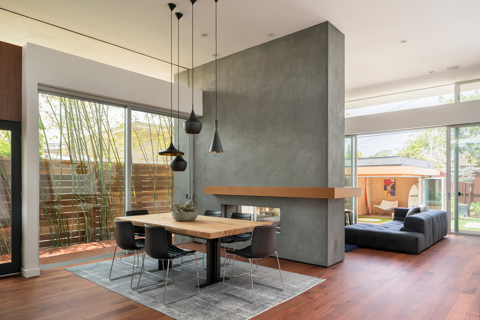 This is an example of a modern dining room in Los Angeles with white walls, dark hardwood floors, a two-sided fireplace, a concrete fireplace surround and brown floor.
