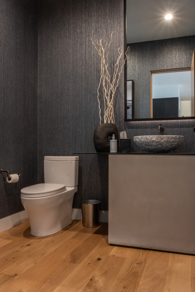 Design ideas for a mid-sized contemporary powder room in Other with flat-panel cabinets, grey cabinets, a two-piece toilet, grey walls, medium hardwood floors, a vessel sink, engineered quartz benchtops, brown floor, black benchtops, a built-in vanity and wallpaper.
