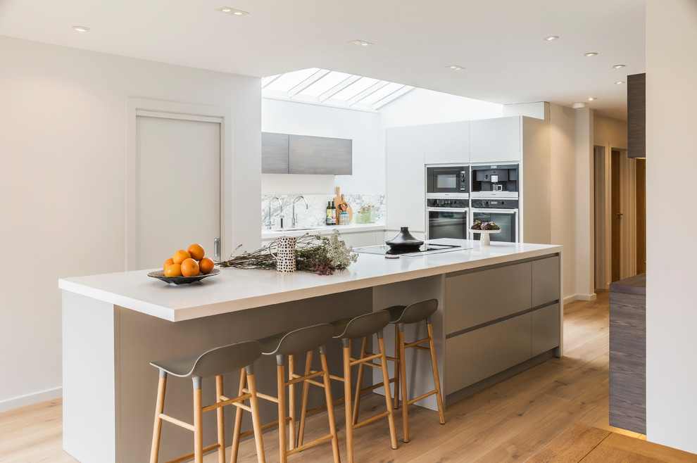 Photo of a contemporary u-shaped open plan kitchen in London with flat-panel cabinets, stainless steel appliances, medium hardwood floors, with island, brown floor, white benchtop, a drop-in sink, grey cabinets, solid surface benchtops, white splashback and marble splashback.
