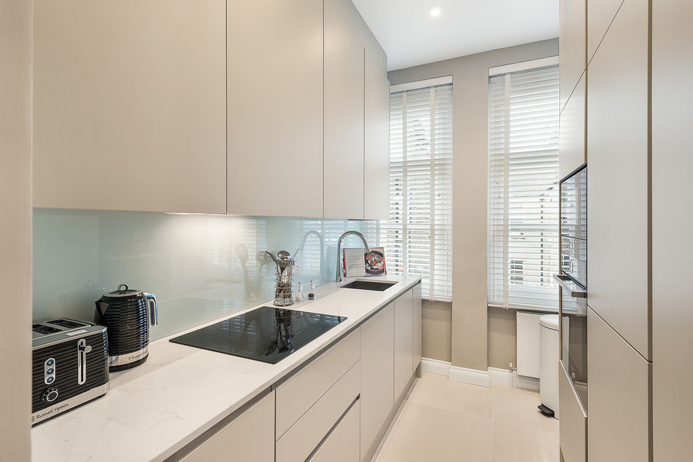 Small contemporary galley separate kitchen in Essex with a drop-in sink, flat-panel cabinets, beige cabinets, marble benchtops, blue splashback, glass sheet splashback, black appliances, ceramic floors, no island, beige floor and white benchtop.