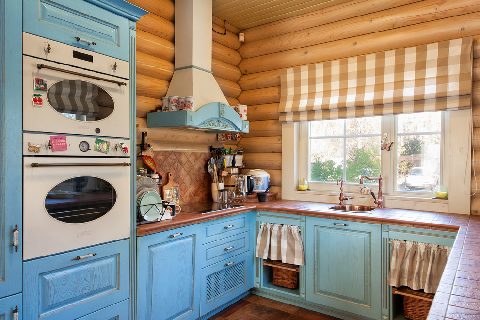 Photo of a mid-sized country u-shaped eat-in kitchen in Saint Petersburg with a drop-in sink, raised-panel cabinets, blue cabinets, tile benchtops, brown splashback, white appliances and brown floor.