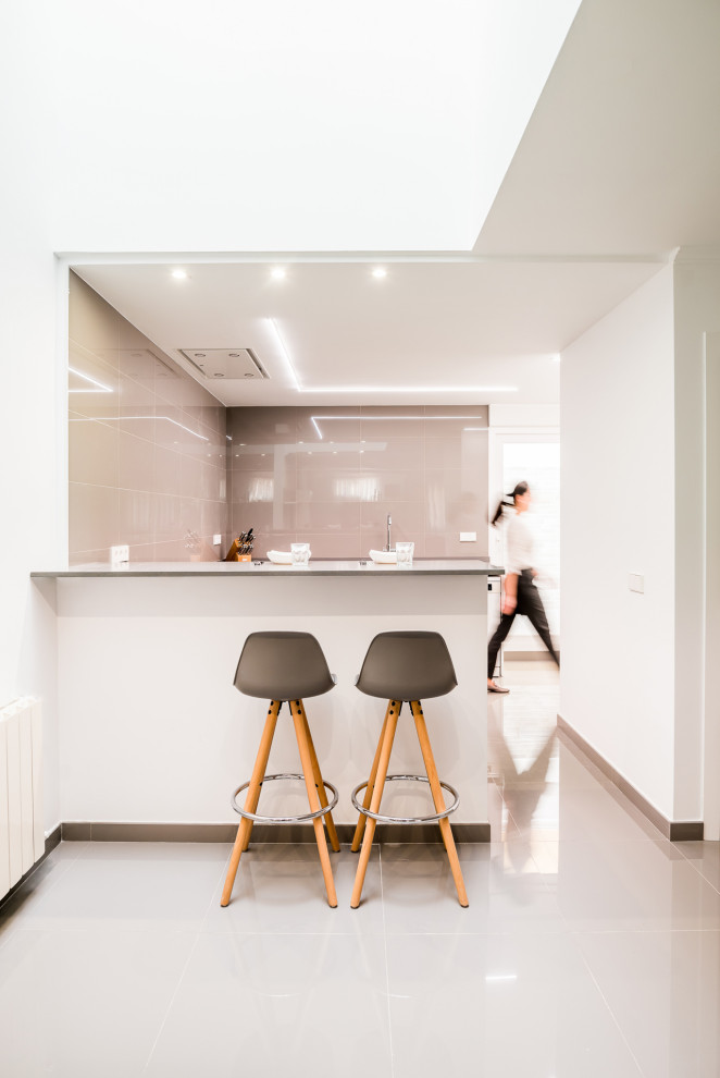 Mid-sized contemporary u-shaped open plan kitchen in Valencia with an undermount sink, flat-panel cabinets, white cabinets, quartz benchtops, grey splashback, porcelain splashback, white appliances, porcelain floors, a peninsula, grey floor and grey benchtop.