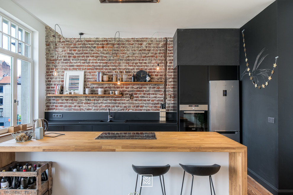 Photo of a small industrial galley separate kitchen in Lyon with a drop-in sink, flat-panel cabinets, black cabinets, wood benchtops, red splashback, brick splashback, stainless steel appliances, medium hardwood floors, a peninsula and brown floor.