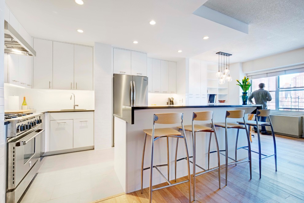Photo of a small contemporary l-shaped open plan kitchen in New York with a single-bowl sink, flat-panel cabinets, white cabinets, quartz benchtops, white splashback, subway tile splashback, stainless steel appliances, porcelain floors, with island, grey floor and grey benchtop.
