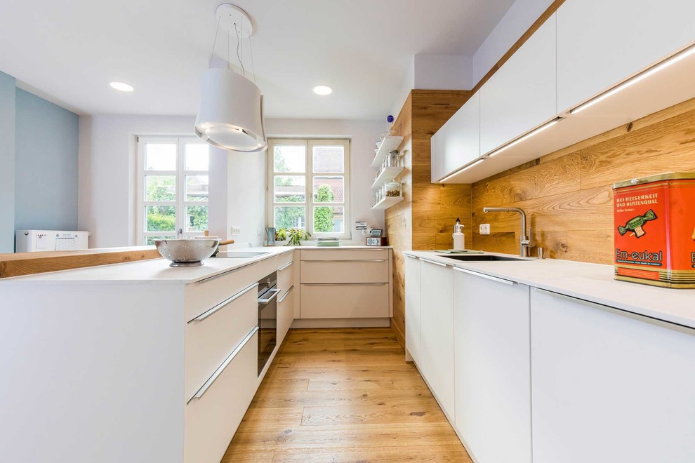 Photo of a mid-sized scandinavian u-shaped separate kitchen in Nuremberg with an integrated sink, flat-panel cabinets, white cabinets, brown splashback, timber splashback, black appliances, medium hardwood floors, a peninsula, brown floor and white benchtop.