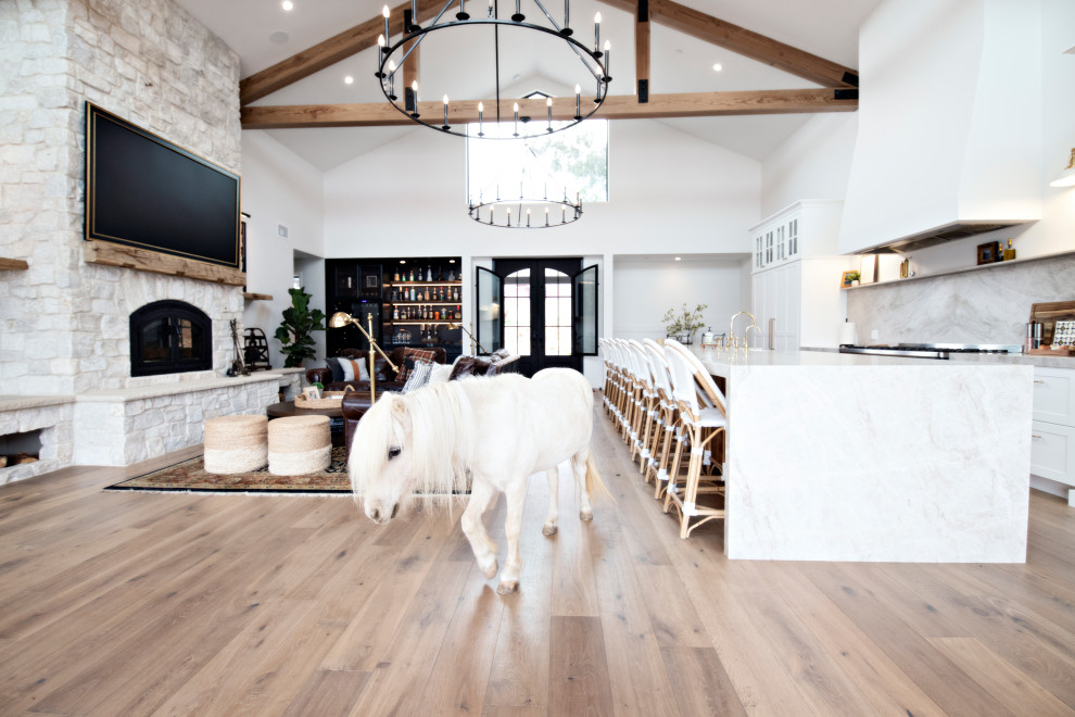 Inspiration for a large farmhouse single-wall light wood floor, beige floor and exposed beam open concept kitchen remodel in San Diego with a farmhouse sink, shaker cabinets, white cabinets, quartzite countertops, white backsplash, marble backsplash, black appliances, an island and white countertops