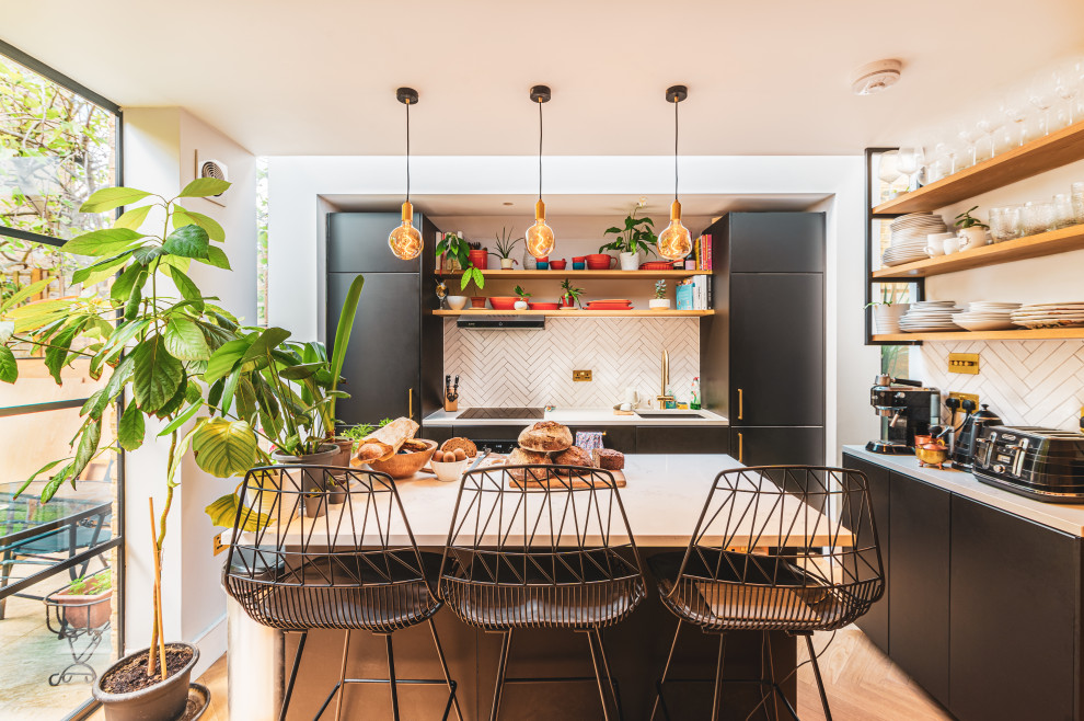Inspiration for a small bohemian single-wall kitchen/diner in London with a built-in sink, flat-panel cabinets, black cabinets, composite countertops, white splashback, porcelain splashback, stainless steel appliances, light hardwood flooring, an island and white worktops.