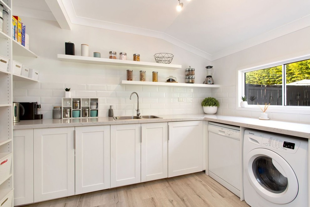 This is an example of a mid-sized l-shaped kitchen pantry in Brisbane with a drop-in sink, recessed-panel cabinets, white cabinets, quartz benchtops, subway tile splashback, black appliances, laminate floors and with island.