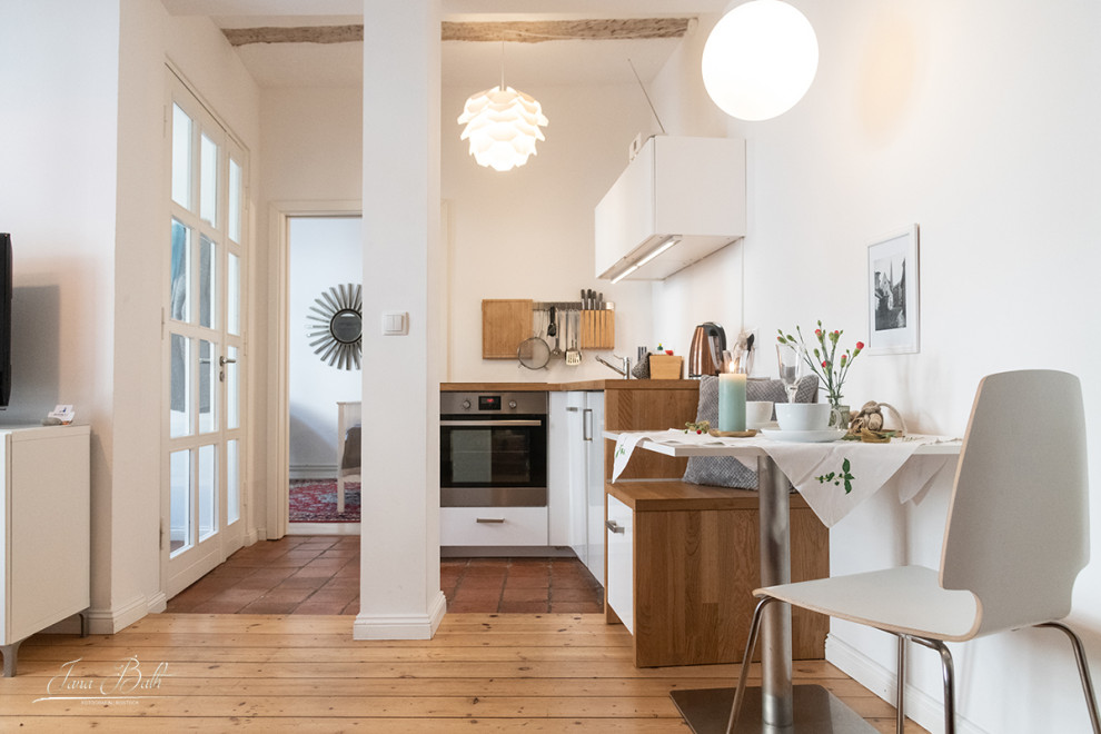 Photo of a small scandinavian l-shaped open plan kitchen in Other with a single-bowl sink, flat-panel cabinets, white cabinets, wood benchtops, metallic splashback, stainless steel appliances, light hardwood floors, no island and brown benchtop.