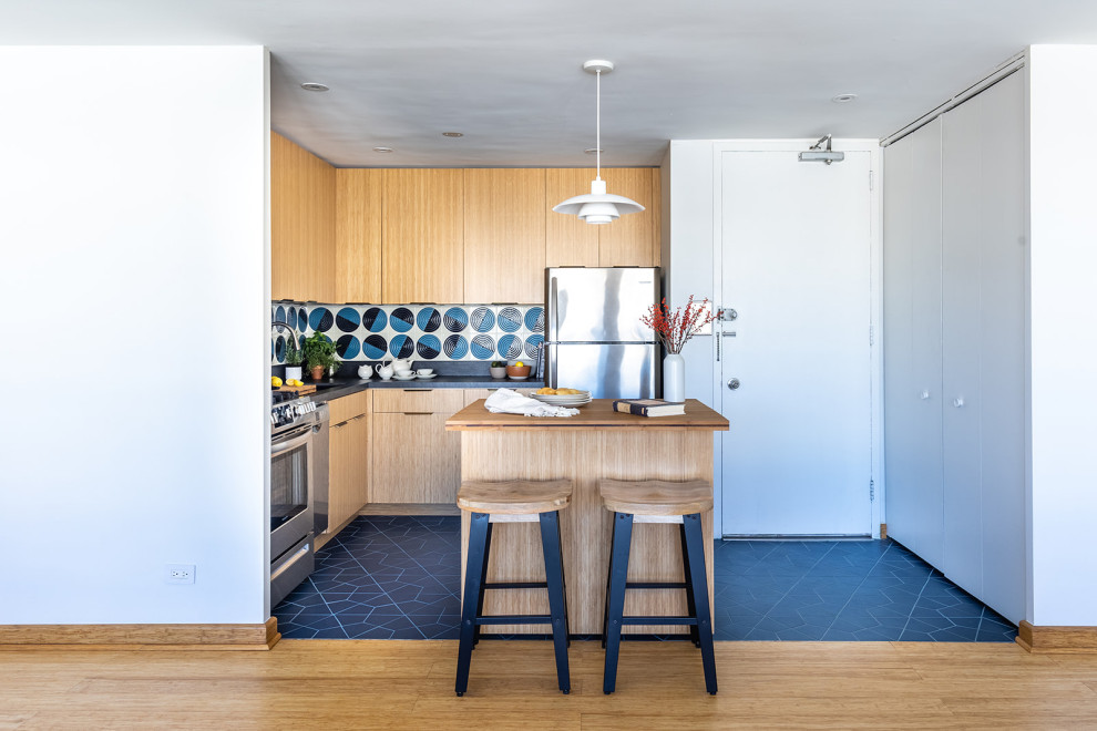 Photo of a small eclectic l-shaped open plan kitchen in Chicago with an undermount sink, flat-panel cabinets, light wood cabinets, solid surface benchtops, blue splashback, ceramic splashback, stainless steel appliances, ceramic floors, with island, blue floor and black benchtop.