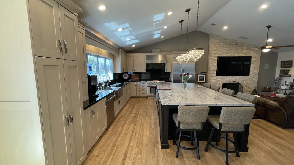 Mid-sized transitional l-shaped kitchen pantry in Orange County with a farmhouse sink, shaker cabinets, white cabinets, granite benchtops, black splashback, granite splashback, stainless steel appliances, light hardwood floors, with island, brown floor, multi-coloured benchtop and coffered.