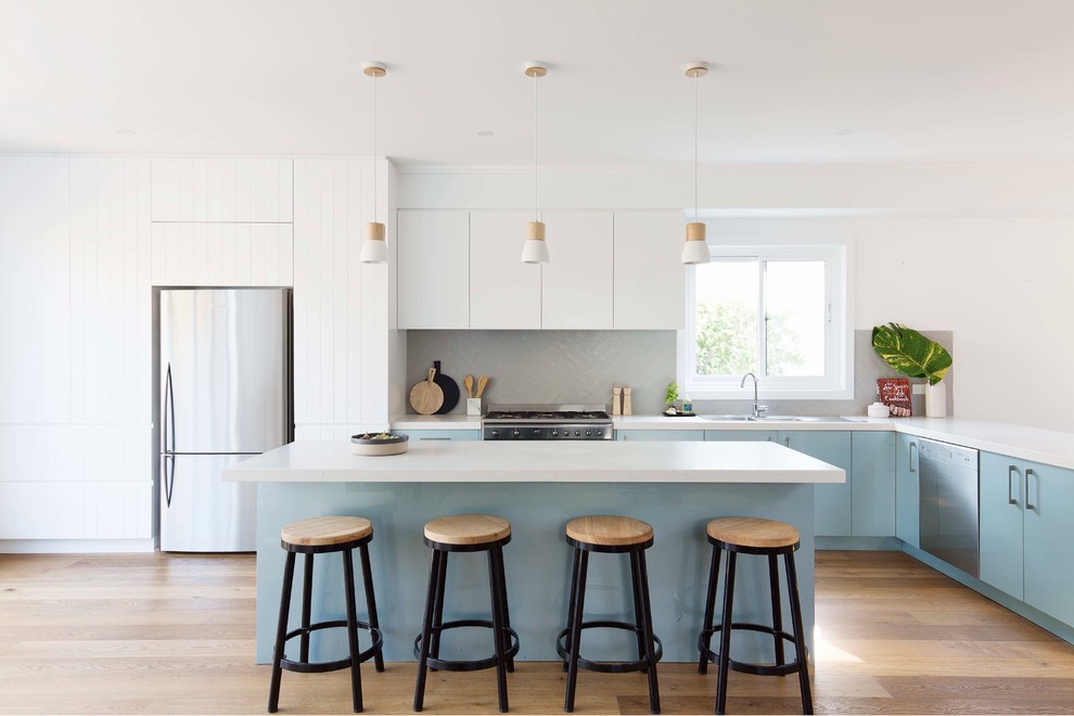 This is an example of a beach style l-shaped kitchen in Sydney with a double-bowl sink, flat-panel cabinets, blue cabinets, grey splashback, stainless steel appliances, medium hardwood floors, with island and brown floor.