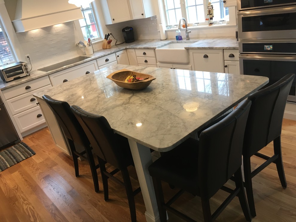 Inspiration for a transitional l-shaped medium tone wood floor eat-in kitchen remodel in Boston with a farmhouse sink, shaker cabinets, white cabinets, white backsplash, porcelain backsplash, stainless steel appliances and an island