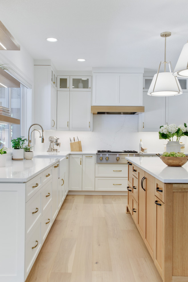 Photo of an expansive classic u-shaped kitchen/diner in Portland with a belfast sink, shaker cabinets, white cabinets, engineered stone countertops, white splashback, stone slab splashback, stainless steel appliances, light hardwood flooring, an island, beige floors and white worktops.