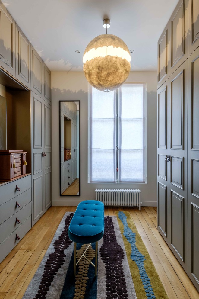 Photo of a large transitional gender-neutral dressing room in Paris with grey cabinets and light hardwood floors.