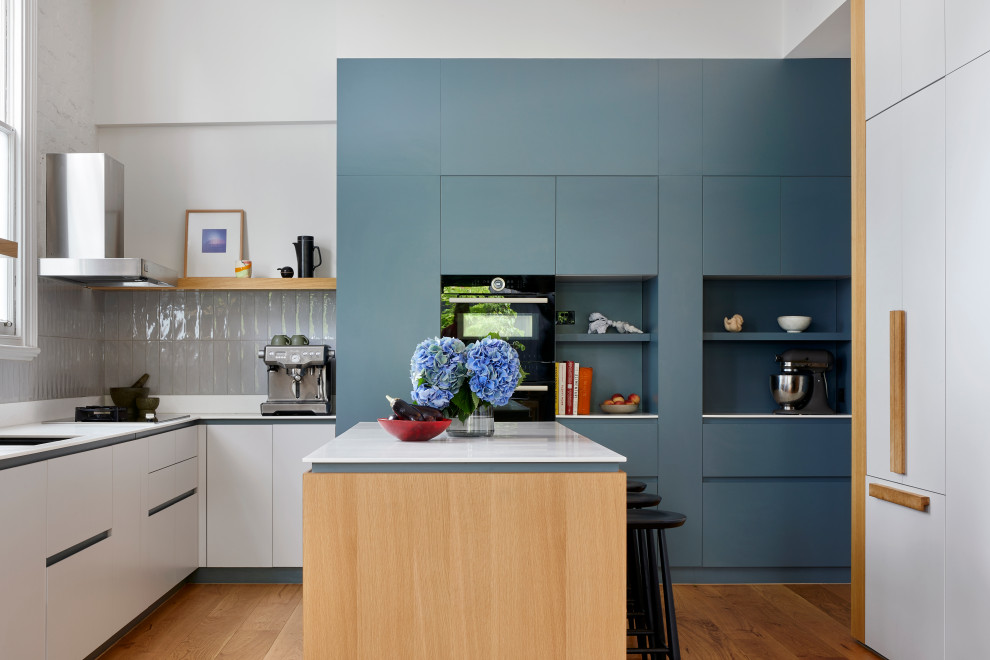 Design ideas for a mid-sized contemporary u-shaped open plan kitchen in Melbourne with an undermount sink, flat-panel cabinets, blue cabinets, solid surface benchtops, grey splashback, ceramic splashback, black appliances, medium hardwood floors, with island, beige floor and white benchtop.
