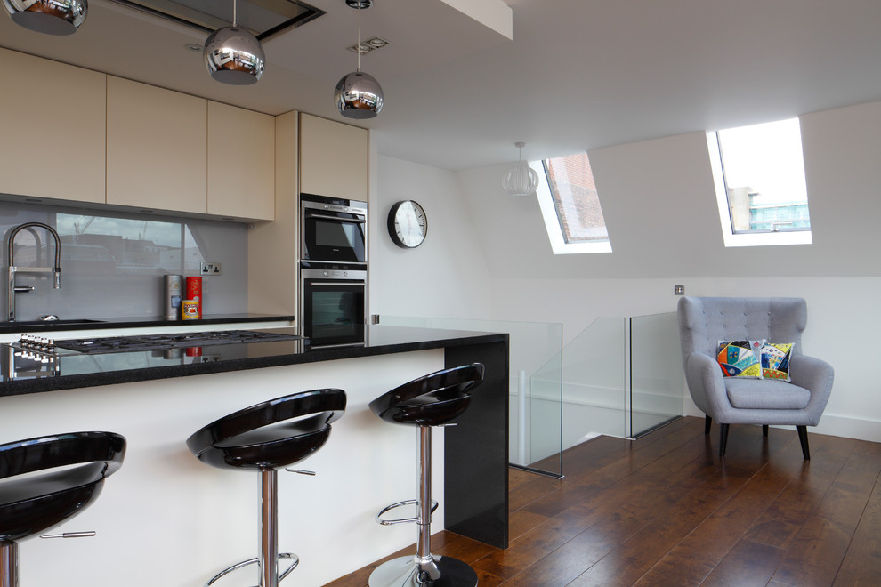 Large contemporary l-shaped open plan kitchen in London with grey splashback, medium hardwood floors, with island, an integrated sink, flat-panel cabinets, white cabinets, granite benchtops, glass sheet splashback, panelled appliances, brown floor and black benchtop.