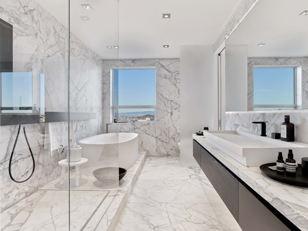 This is an example of a large contemporary master bathroom in San Francisco with flat-panel cabinets, dark wood cabinets, a freestanding tub, a corner shower, marble, white walls, marble floors, a trough sink, marble benchtops, white floor, an open shower, a two-piece toilet, white tile and white benchtops.
