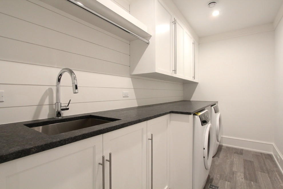 Photo of a mid-sized transitional single-wall dedicated laundry room in New York with an undermount sink, shaker cabinets, white cabinets, white walls, medium hardwood floors, a side-by-side washer and dryer, brown floor and black benchtop.